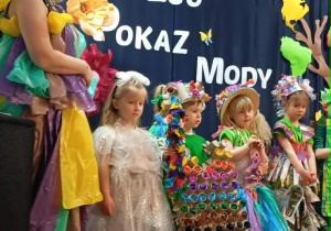 Grupa dzieci w kolorowych strojach ekologicznych stoi na scenie