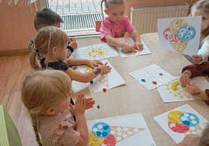 Przedszkolaki siedzą przy stoliku i wykonują pracę plastyczną.