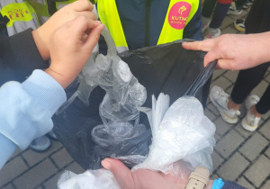 Dzieci w kamizelkach odblaskowych wkładają zebrane odpady do czarnego worka podczas akcji „Sprzątanie Świata”