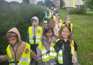Przedszkolaki w kamizelkach odblaskowych idą chodnikiem w kierunku miejsca sprzątania okolicy