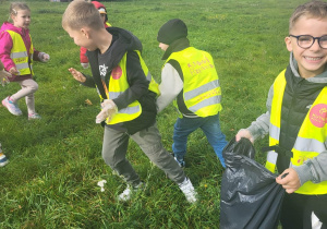 Dzieci w rękawiczkach i kamizelkach odblaskowych zbierają śmieci na trawniku do czarnych worków