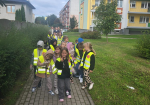 Grupa przedszkolaków w kamizelkach odblaskowych wraca chodnikiem po wspólnym sprzątaniu okolicy