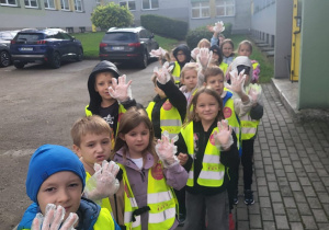 Przedszkolaki stoją przed budynkiem przedszkola i pokazują założone rękawiczki przygotowane do sprzątania