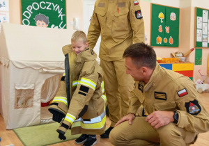 Przedszkolak stoi ubrany w kurtkę strażacką.