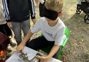 Chłopiec z zasłoniętymi oczami uczestniczy w konkurencji z wykorzystaniem ręcznej maszynki podczas pikniku rodzinnego.