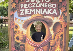 Dziewczynka pozuje w kolorowej fotobudce z ziemniakiem, ozdobionej jesiennymi liśćmi i słonecznikami.