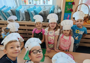 Przedszkolaki w papierowych czapkach kucharskich i kolorowych fartuszkach przygotowują się do zajęć kulinarnych.