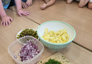 Dzieci oglądają przygotowane składniki sałatki: pokrojone ziemniaki, ogórki, cebulę i świeży koperek.