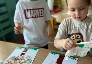 Dwoje dzieci wykonuje prace plastyczne z jesiennym drzewem, odbijając kolorowe wzory na kartkach za pomocą rolek.