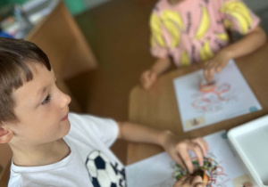 Dzieci podczas zajęć plastycznych odbijają farbę na kartkach z rysunkiem drzewa, tworząc kolorowe jesienne korony.