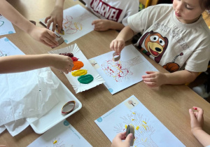 Grupa dzieci siedzi wokół stolika i wspólnie wykonuje jesienne prace plastyczne przedstawiające drzewa z kolorowymi koronami.