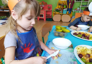 Dziewczynka kroi plastikowym nożykiem owoce do sałatki