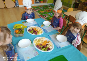 Dzieci siedzą przy stoliku , gdzie będą wykonywać sałatkę owocową