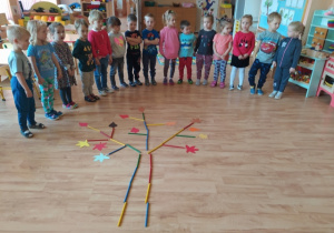 Dzieci stoją wokól sylwety drzewa ułożonego na podłodze z drewnianych patyczków